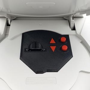 Puede incluir: Consola blanca abierta, que revela un panel de control negro con botones direccionales naranjas y botones de acción rojos. El texto "RFW RETROFUTURE WORKS" en rosa está impreso encima del panel de control.