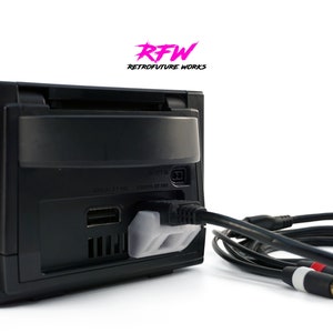 Könnte beinhalten: Schwarze Videospielkonsole mit angeschlossenen Kabeln. Die Konsole hat einen Schlitz für eine Disc und verschiedene Anschlüsse. Der Text "RFW RETROFUTURE WORKS" ist oben links zu sehen.