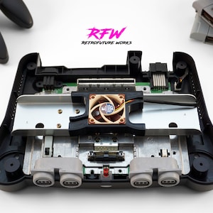 May include: A black Nintendo 64 console with the top removed, revealing the internal components. A small fan is installed on the motherboard, and the console has two controller ports. The text "RFW RETROFUTURE WORKS" is visible in the background.