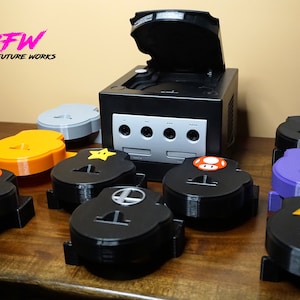 May include: A black GameCube console with several custom-designed controller port covers. The covers are black, orange, gray, and purple, each featuring a different video game icon, including a star, mushroom, and Triforce symbol.