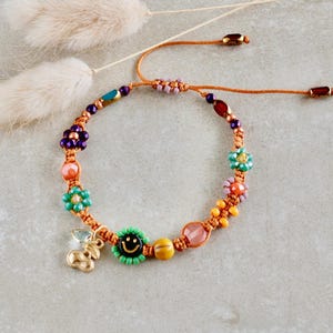 Puede incluir: Una colorida pulsera de cuentas con diseño macramé. La pulsera presenta varias cuentas con forma de flor en tonos morado, turquesa y naranja, junto con un dije dorado y una cuenta verde con una carita sonriente. Fondo neutro.