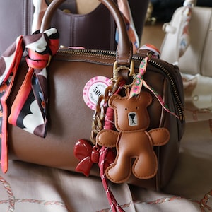 Może przedstawiać: Brązowa skórzana torebka z górnym uchwytem i zapięciem na zamek błyskawiczny. Torba jest ozdobiona kolorową chustą, breloczkiem do kluczy w kształcie misia i czerwoną kokardą. Inne torebki są widoczne w tle.
