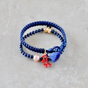 Op de afbeelding: Een dubbelgelaagde armband met donkerblauw gevlochten koord en kleine donkerblauwe kralen. Het sieraad bevat een enkele lichtroze parel, een kleine rode koraalvormige bedel en een klein blauw kwastje, afgewerkt met goudkleurige metalen details.