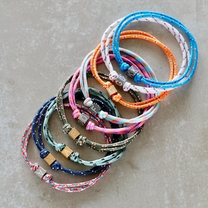 May include: A collection of colorful rope bracelets with wooden letter beads. The bracelets are in various colors, including blue, orange, pink, and green. Each bracelet has a different letter or combination of letters on the wooden beads.
