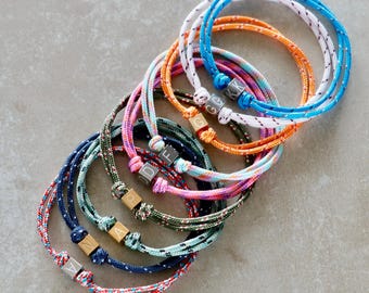 May include: A collection of colorful rope bracelets with wooden letter beads. The bracelets are in various colors, including blue, orange, pink, and green. Each bracelet has a different letter or combination of letters on the wooden beads.