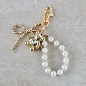 Peut inclure: Un porte-clés doré avec une chaîne de perles, un petit charme en forme d'ours avec un strass et un charme en forme de nœud doré.