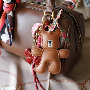 Może przedstawiać: Brązowa skórzana torebka z złotym zamkiem i uchwytem, ozdobiona brelokiem w kształcie misia. Brelok jest brązowy z haftowanymi detalami i czerwono-białym sznurkiem. Wzór szalika jest zawiązany na uchwycie.