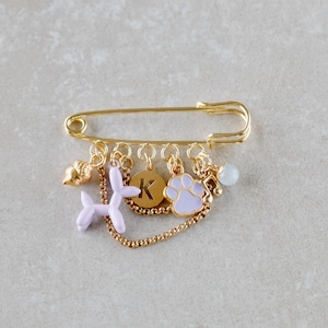 Könnte beinhalten: Eine goldfarbene Sicherheitsnadel mit mehreren Anhängern. Zu den Anhängern gehören ein Ballonhund, ein Pfotenabdruck, eine Scheibe mit dem Buchstaben "K" und eine kleine Muschel. Der Pfotenabdruck und der Ballonhund sind hellviolett.