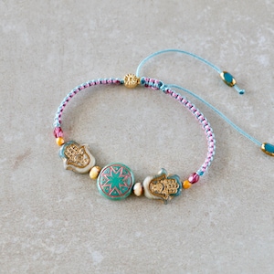 Könnte beinhalten: Ein geflochtenes Armband mit einer zentralen, runden, blaugrünen Perle mit kupferfarbenem Sternmuster, flankiert von zwei goldfarbenen Hamsa-Hand-Anhängern. Das Band besteht aus hellblauem und rosafarbenem Kordelmaterial mit kleinen gelben und rosa Perlen.