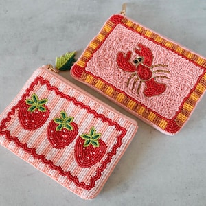 Puede incluir: Dos estuches rectangulares con cremallera y abalorios detallados. Uno es rosa claro con tres fresas rojas y un tirador de cremallera en forma de hoja verde. El otro es rosa claro con un motivo de langosta roja y un borde a cuadros amarillo y rojo.