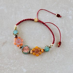 Könnte beinhalten: Ein Armband mit einer dunkelroten geflochtenen Kordel und einem verstellbaren Schiebeverschluss. Das Design umfasst mehrere blumenförmige Perlen in Orange-, Gelb- und Türkistönen, akzentuiert durch kleine goldene Metallperlen und eine einzelne rote Perle.
