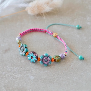 Op de afbeelding: Een armband met een gevlochten roze koord en verstelbare turquoise touwtjes. Het ontwerp bevat keramische bloemkralen in tinten blauwgroen, paars en groen, geaccentueerd door kleine oranje en goudkleurige kralen langs de band.