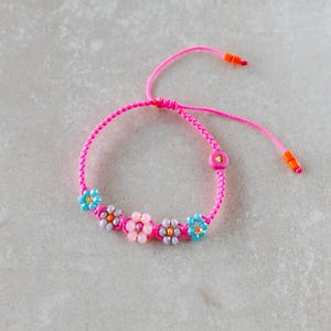 May include: A bright pink braided cord bracelet featuring five flower-shaped bead clusters in shades of light blue, grey, and pale pink. The bracelet includes a pink circular bead with a small gold heart and two orange cylindrical end beads.