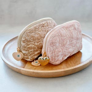 Könnte beinhalten: Zwei Kosmetiktaschen aus Samt in Beige und Rosa, mit Steppmuster und goldenen Reißverschlüssen. Jede Tasche hat einen herzförmigen Anhänger mit einem Augendesign oder dem Wort "LOVE". Die Taschen liegen auf einem Holztablett.