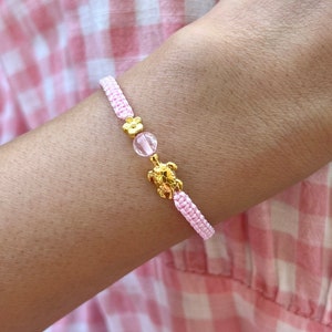 Könnte beinhalten: Ein pinkfarbenes geflochtenes Armband mit einem goldenen Schildkrötenanhänger und einer rosafarbenen Kristallperle.