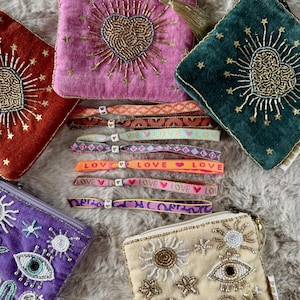 May include: A collection of velvet pouches featuring beaded heart and eye motifs alongside several fabric wristbands with woven patterns and the word LOVE printed on them, each secured with a small white heart-shaped bead closure.