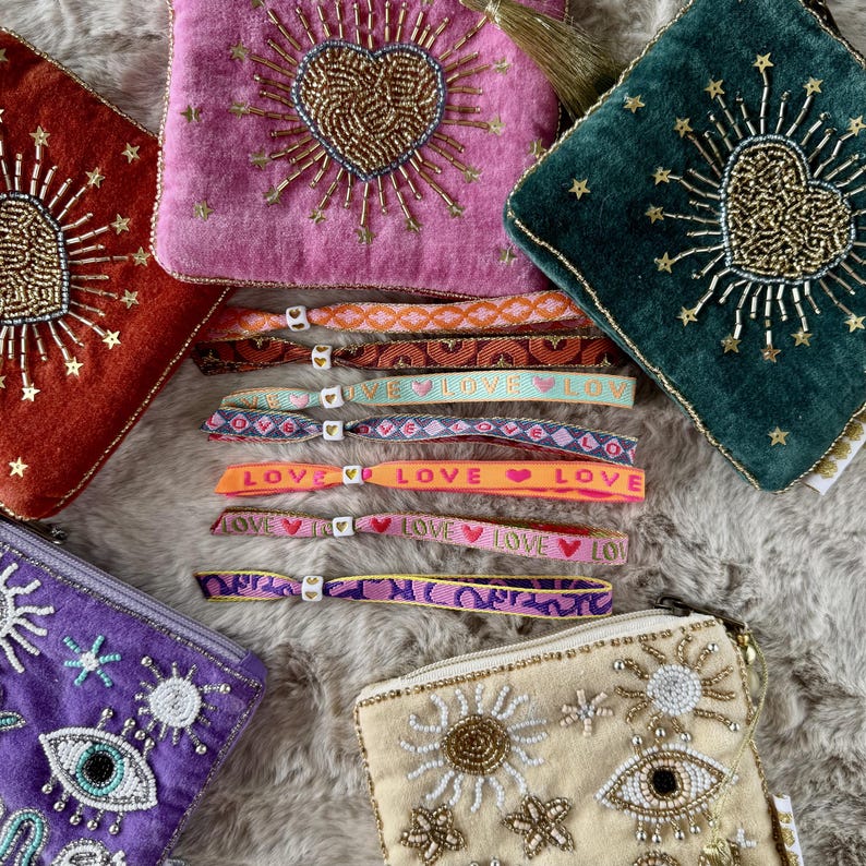 May include: A collection of velvet pouches featuring beaded heart and eye motifs alongside several fabric wristbands with woven patterns and the word LOVE printed on them, each secured with a small white heart-shaped bead closure.