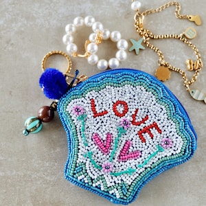 Könnte beinhalten: Eine Perlenbörse mit dem Wort "LOVE" in Rot, umgeben von rosa Blumen und grünen Stielen. Die Börse ist blau, weiß und grün. Ein blauer Pompon-Schlüsselanhänger und Perlenschmuck sind ebenfalls sichtbar.