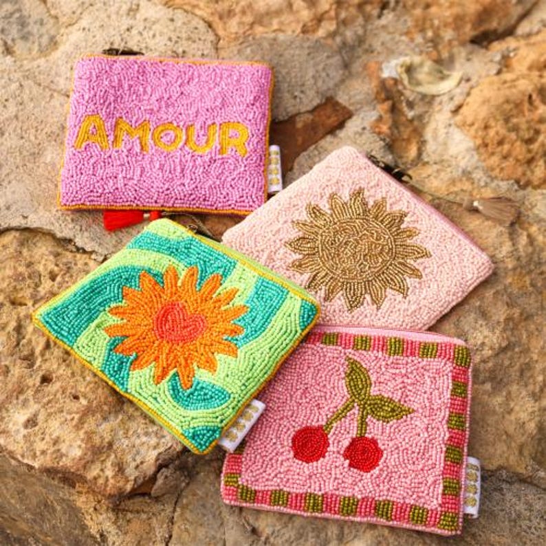 May include: Four square zipped pouches covered in small beads. One is pink with the yellow text AMOUR. One is light pink with a gold beaded sun. One is bright green and teal with an orange flower. One is pink with a red cherry design.