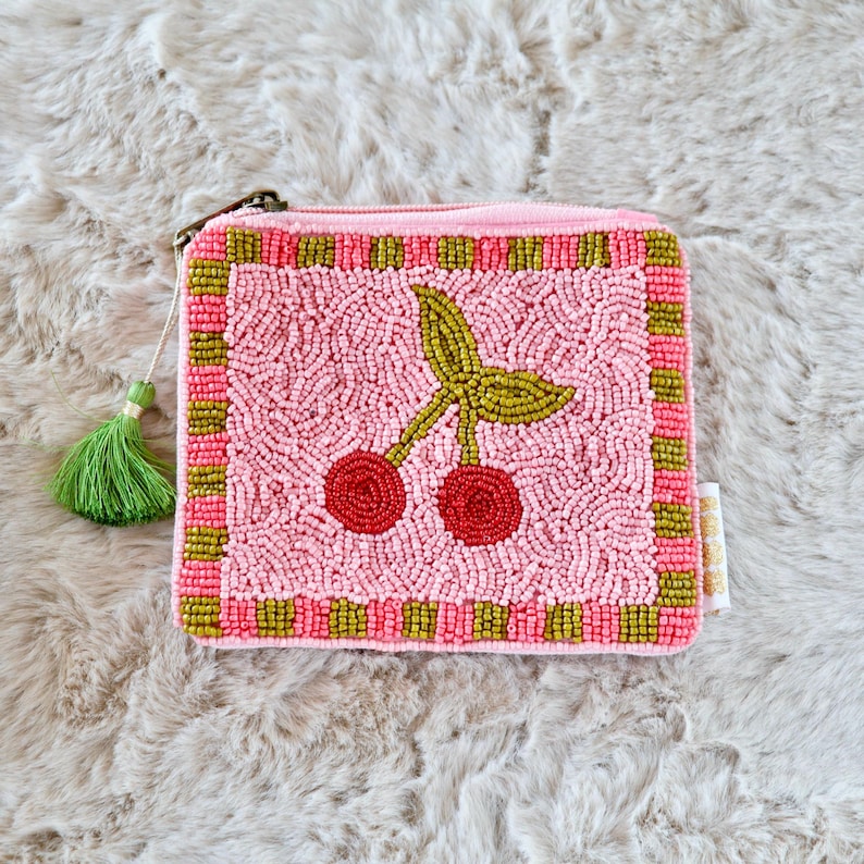 May include: A square zipped pouch featuring a beaded cherry design in red and green on a light pink background. The border consists of alternating pink and olive green beaded squares. A green tassel is attached to the metal zip pull.