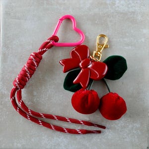 Könnte beinhalten: Ein rot-weißes Schlüsselband mit einem herzförmigen rosa Clip und einem goldfarbenen Verschluss. Der Anhänger zeigt eine rote Schleife, grüne Blätter und zwei rote Samtkirschen. Ein dekoratives Accessoire.