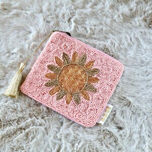 May include: A square coin purse covered in light pink beads with a central sunflower design made of gold and bronze beads. The purse features a gold-coloured metal zipper pull with a matching gold tassel attached to the side and a small white tag.