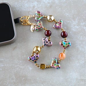 Può includere: Un ciondolo per telefono con perline a forma di cuore con un motivo leopardato. Il ciondolo presenta perline e hardware color oro, con un cinturino nero per l'attacco. Il design include perline di accento rosse e arancioni.