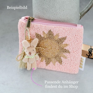 May include: A light pink beaded coin purse featuring a gold beaded sun design on the front. A gold zip closure is attached to a gold chain with a small, light beige plush rabbit charm and a light beige tassel hanging from the side.