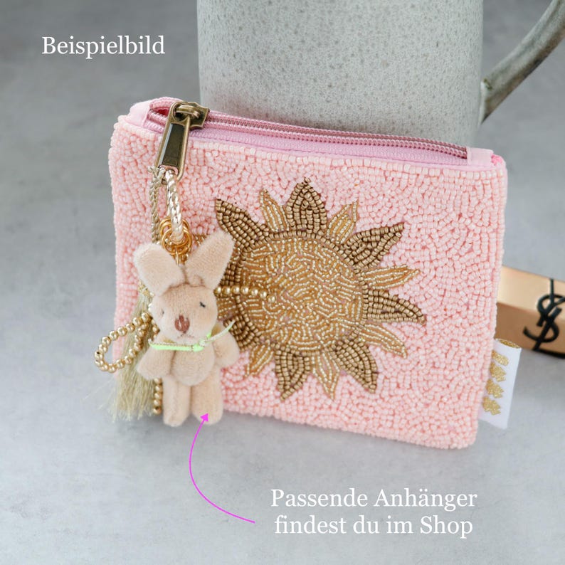 May include: A light pink beaded coin purse featuring a gold beaded sun design on the front. A gold zip closure is attached to a gold chain with a small, light beige plush rabbit charm and a light beige tassel hanging from the side.