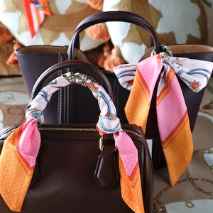 Könnte beinhalten: Zwei braune Lederhandtaschen mit dekorativen Seidenschals. Eine Tasche hat einen Schal um den Griff gewickelt, die andere ist am Griff befestigt. Die Schals zeigen rosa, orange und weiße Muster.