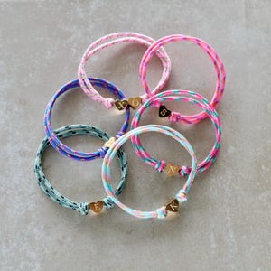 Könnte beinhalten: Eine Sammlung von fünf kreisförmigen Kordelarmbändern in verschiedenen Farben wie Pink, Blau und Türkis. Jedes Armband hat einen kleinen goldfarbenen herzförmigen Anhänger, der mit einem einzelnen Buchstaben graviert ist, nämlich E, K, N, O und X.