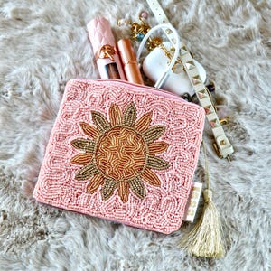 May include: A square pink beaded coin purse featuring a central gold and bronze beaded sunflower design, accompanied by a gold tassel. Included are a pink lipstick, a bronze lipstick, a white wireless earbud case, and a white studded strap.