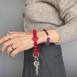 Könnte beinhalten: Ein roter Perlen-Schlüsselanhänger mit herzförmigem Akzent und silbernem Schlüsselring. Der Schlüsselanhänger ist an einem Schlüssel befestigt. Ein rotes Perlenarmband und ein lila Perlenarmband sind ebenfalls sichtbar.
