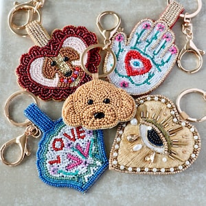 May include: A collection of five beaded keychains featuring diverse designs including a brown dog face, a red heart-shaped dog portrait, a white hamsa hand with a red eye, a blue shape with the word LOVE, and a gold heart with an eye motif.