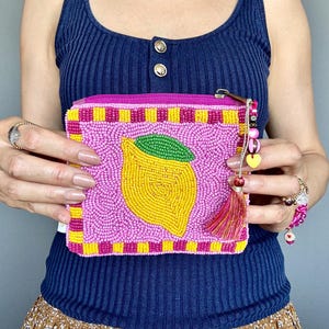 May include: A rectangular zipped pouch covered in pink, yellow, and green seed beads. The front features a central yellow lemon design with a green leaf, surrounded by a chequered border. A decorative tassel and colourful beads hang from the zip.