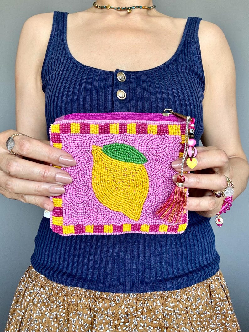 May include: A rectangular zipped pouch covered in pink, yellow, and green seed beads. The front features a central yellow lemon design with a green leaf, surrounded by a chequered border. A decorative tassel and colourful beads hang from the zip.