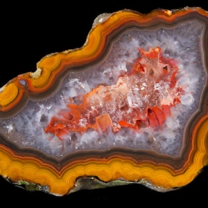 May include: A polished agate slice featuring concentric bands of orange, yellow, and dark brown surrounding a central cavity filled with translucent white quartz crystals and vibrant red mineral deposits against a black background.