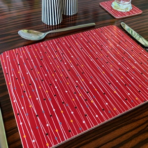 May include: A rectangular placemat featuring a deep red background with a pattern of thin, vertical white lines interspersed with small, colorful square accents in black, white, orange, and blue, arranged in a repeating, linear geometric design.