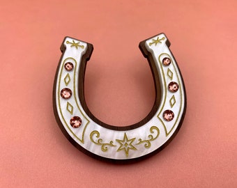 May include: A white and brown horseshoe-shaped brooch with a gold star and floral design. The horseshoe is decorated with pink rhinestones.