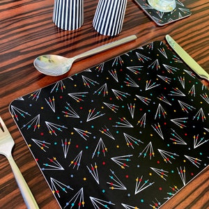 May include: Black placemat with a white geometric pattern and colourful dots. The placemat is on a wooden table with a fork, spoon, and knife.