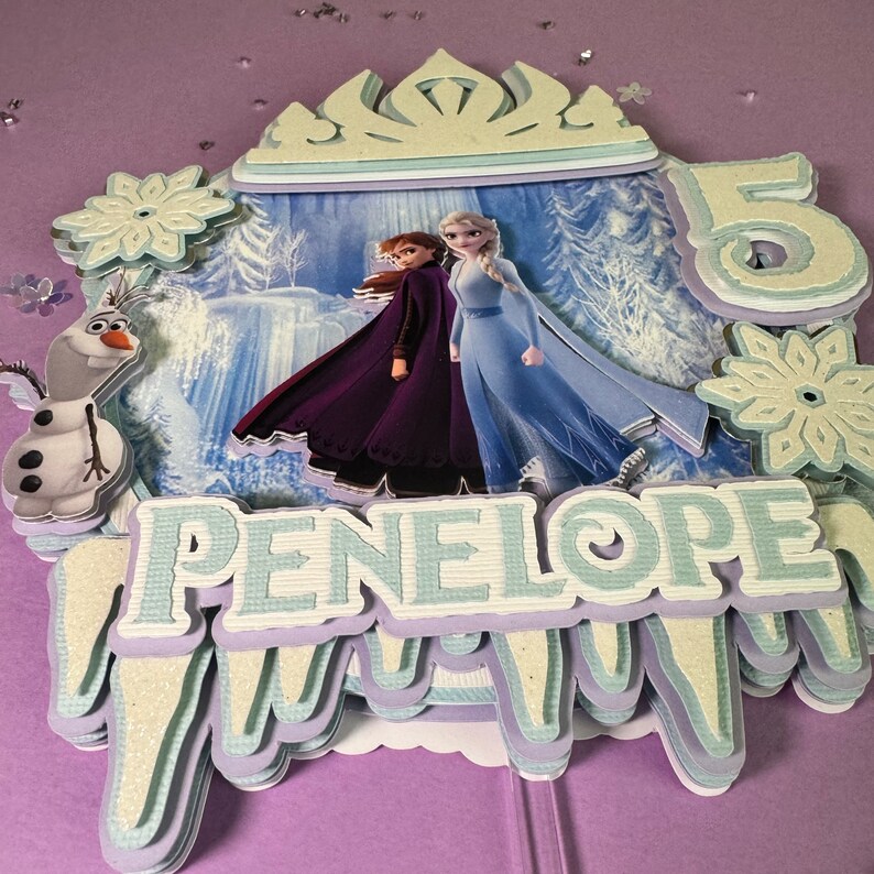 Pode incluir: Um topo de bolo azul e branco com os personagens Elsa e Anna do filme Frozen. O topo tamb&eacute;m inclui uma coroa, flocos de neve e o nome Penelope. O n&uacute;mero 5 tamb&eacute;m est&aacute; inclu&iacute;do no topo.