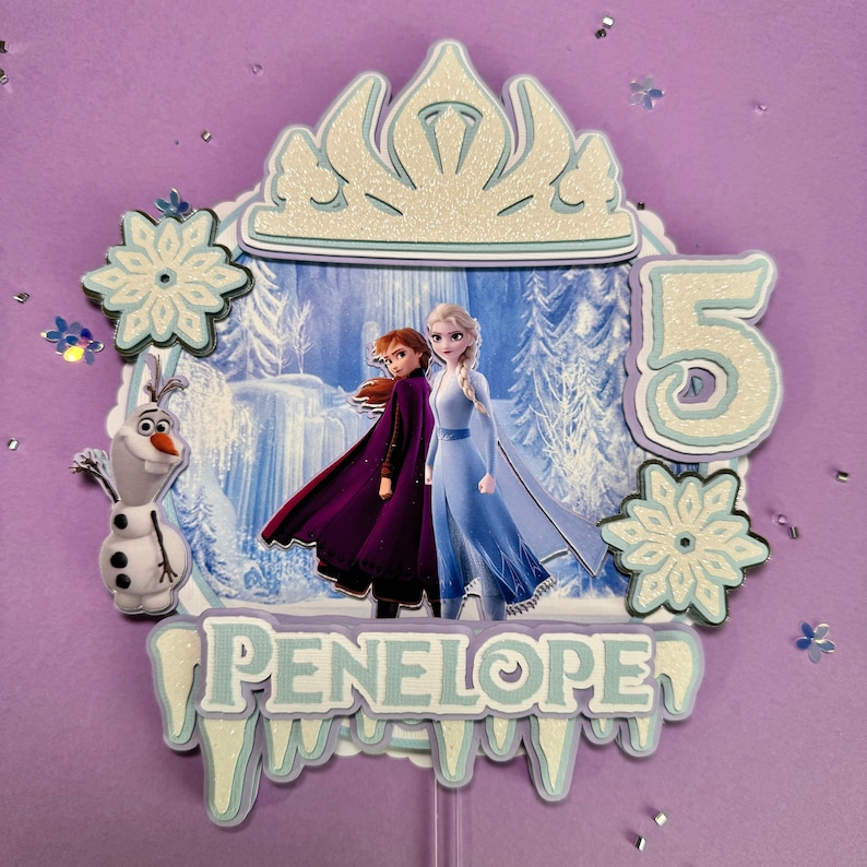 Pode incluir: Um topo de bolo com tema Frozen com Elsa e Anna do filme Frozen. O topo inclui uma coroa branca, um n&uacute;mero 5 azul e branco, duas flocos de neve brancos, uma imagem de Elsa e Anna em azul e branco, e um design de pingentes de gelo azul e branco com o nome Penelope.