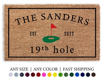May include: A brown coir doormat with black text that reads "THE SANDERS EST. 2022 19th hole". A green and red golf flag is centred on the mat.