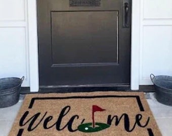 May include: A brown welcome mat with a black border and white cursive text that says "welcome". A green golf course with a red flag is in the middle of the text. The mat is in front of a black door with a white wreath on it.