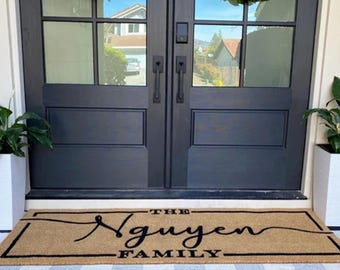 May include: A rectangular tan coir doormat featuring a black border and the black text "THE Nguyen FAMILY" in a script font, layered over a black and white buffalo check rug in front of double dark gray doors with glass panes and green wreaths.