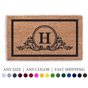 Puede incluir: Una alfombra de puerta rectangular con una superficie de fibra de coco marrón y un borde negro. El centro presenta un diseño circular con la letra "H" y adornos decorativos. El texto "Doormat Decor" está en la parte superior.