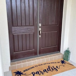 May include: A brown coir doormat with the text "Welcome to our little piece of paradise" in blue cursive lettering with palm tree silhouettes.