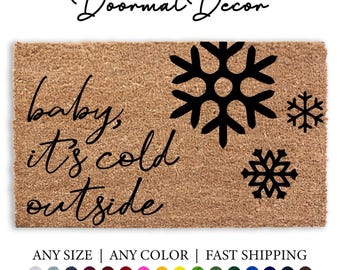 Op de afbeelding: Een bruine kokosvezel deurmat met de tekst "baby, it's cold outside" in zwarte cursieve letters. De deurmat is versierd met zwarte sneeuwvlokontwerpen.
