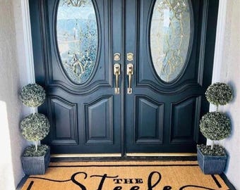 May include: A brown coir doormat with black text that reads "The Steele Family". The doormat is placed in front of a double black door with oval glass panels. Two potted topiary trees are on either side of the door.