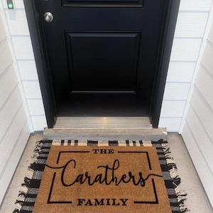 May include: A rectangular doormat with the text "THE Carathers FAMILY" in black script and block letters. The mat is brown with a black and white buffalo plaid border and fringe. The mat is placed in front of a black door.